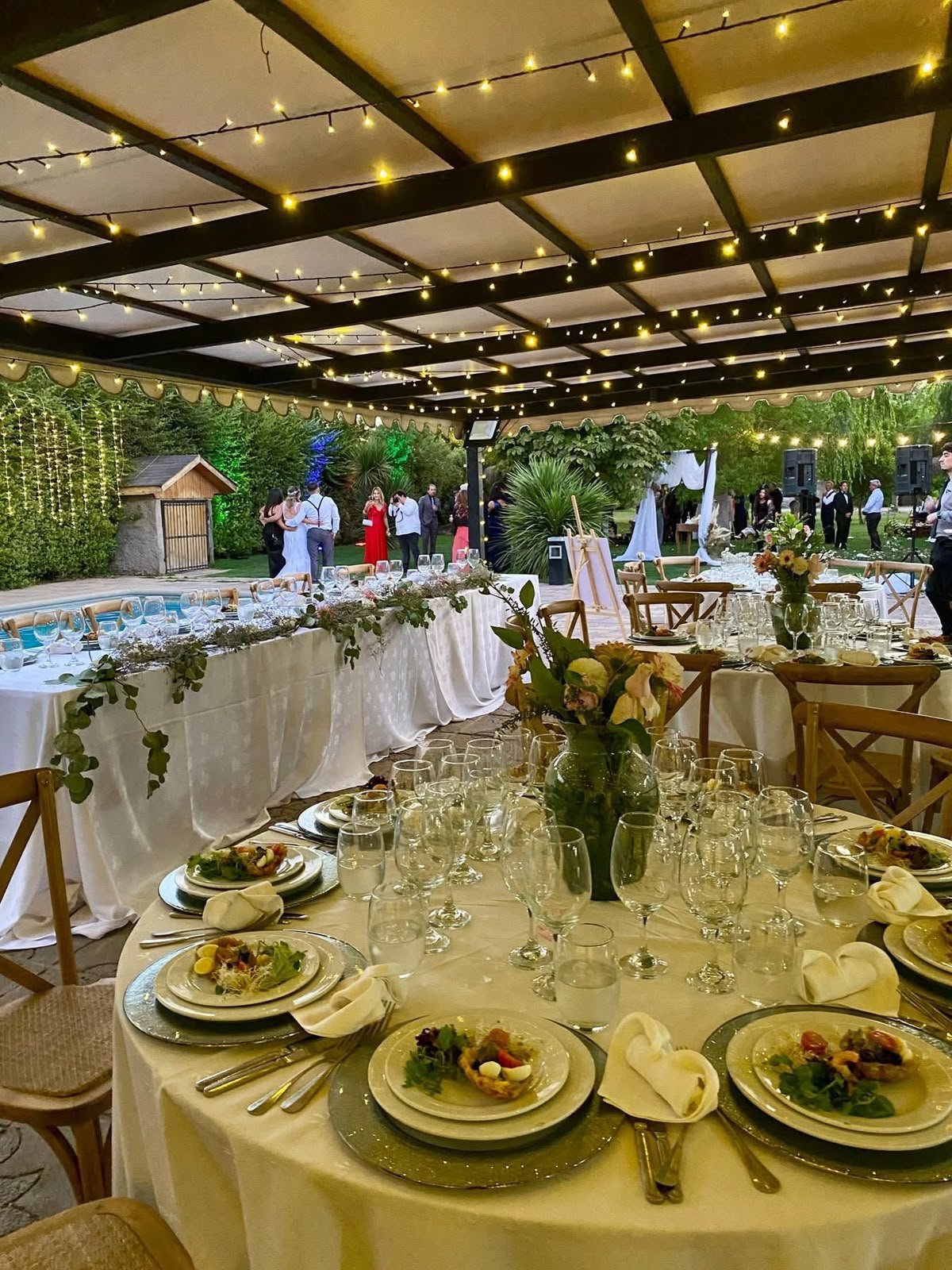 Salón abierto bajo pérgola con guirnaldas de luces, mesas montadas y la piscina al lado